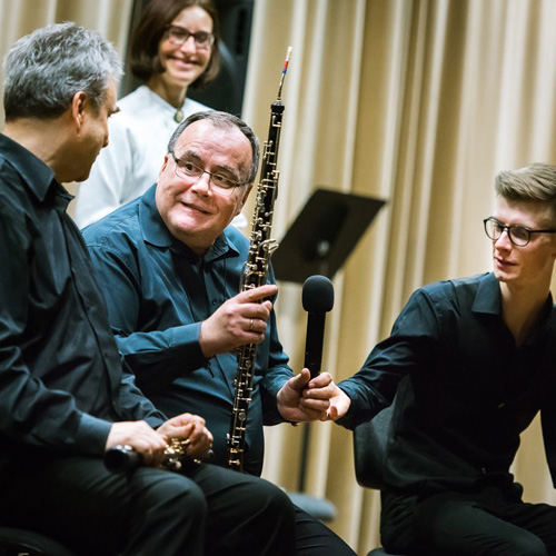trzech muzyków po koncercie, jeden z nich trzyma obój i mikrofon w ręku, w tle prowadząca koncert