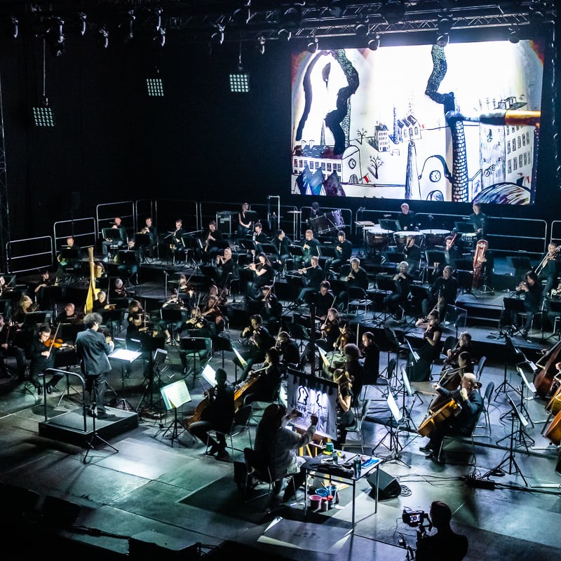 Muzycy orkiestry na scenie wraz z dyrygentem, za nimi ekran z animacją